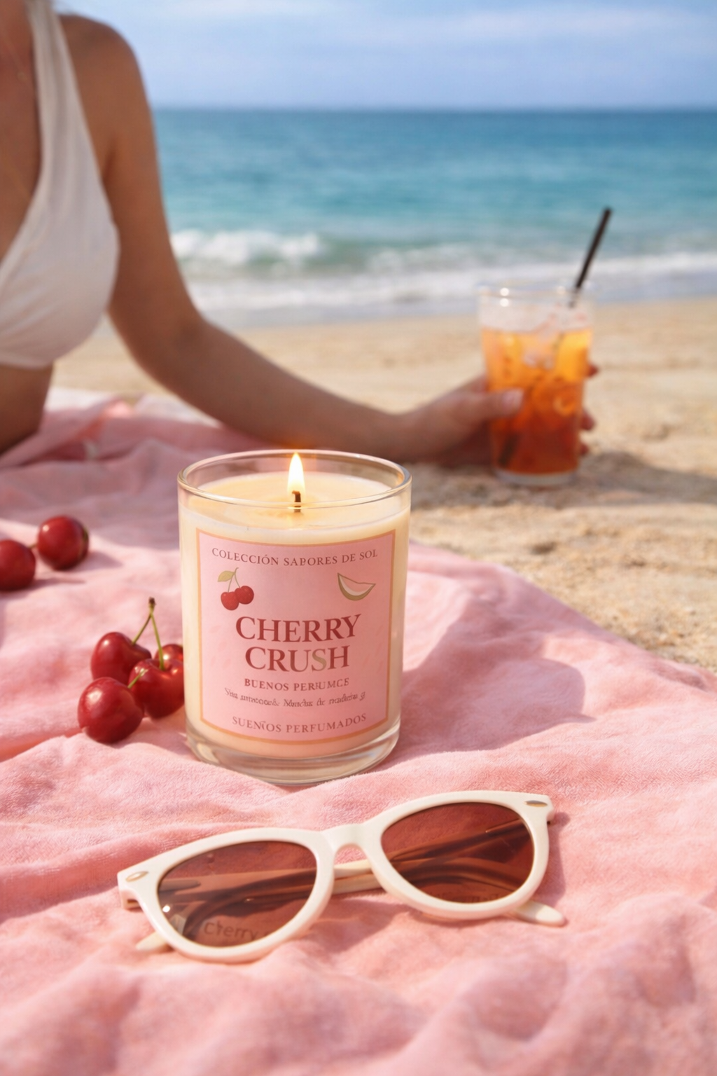 Vela Cherry Crush encendida sobre toalla en la playa, con cerezas frescas y ambiente veraniego